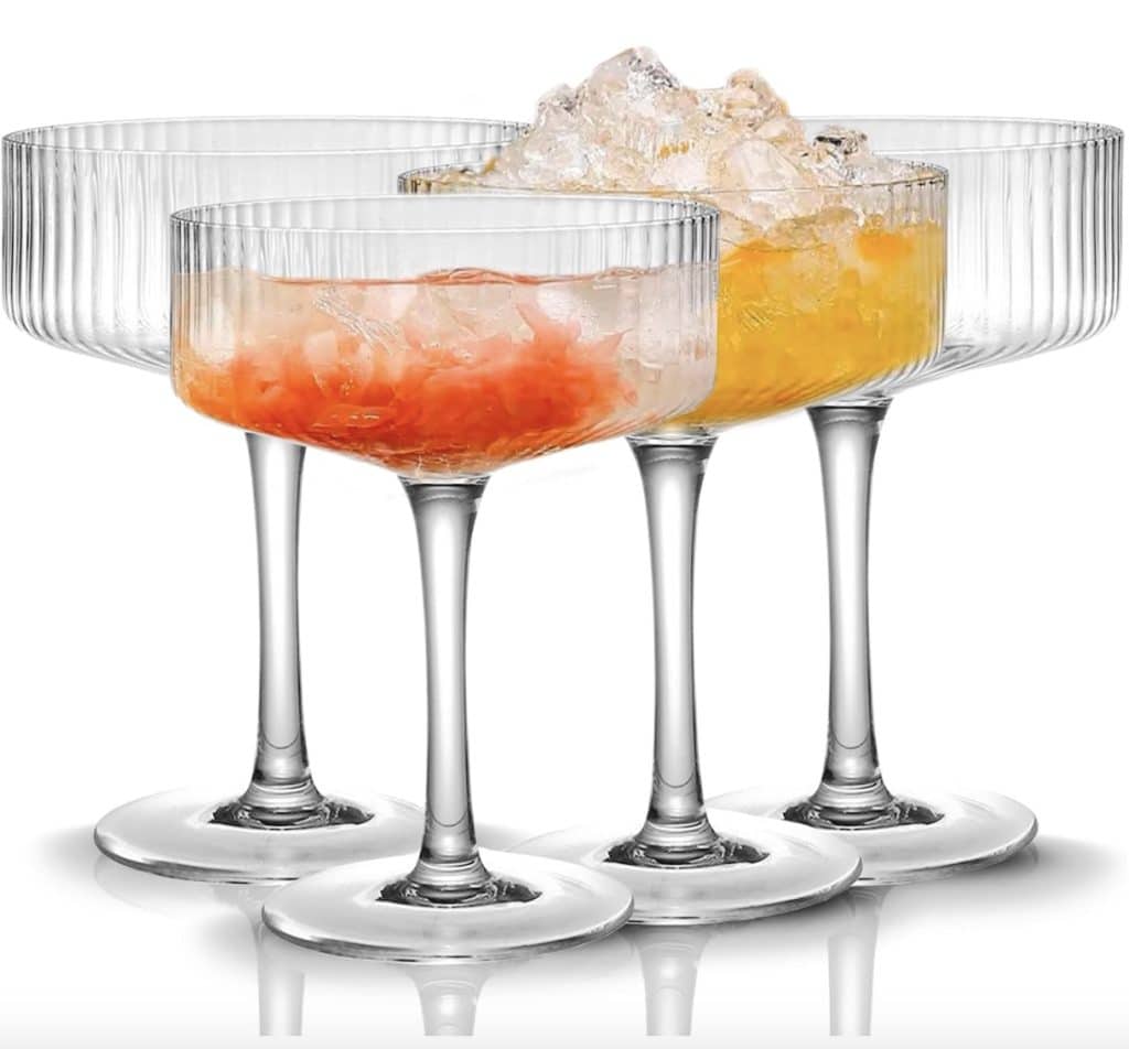 Four elegant cocktail glasses with ribbed designs are filled with crushed ice. Two, perfect for easy summer mocktail recipes, contain orange and red drinks, while the other two remain empty. The glasses have tall stems and clear bases.